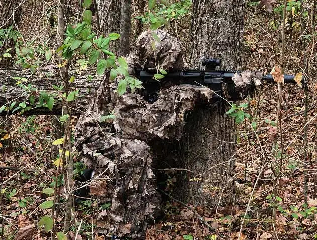Camouflage ghillie suit with leaf and branch patterns, worn by a person in the woods