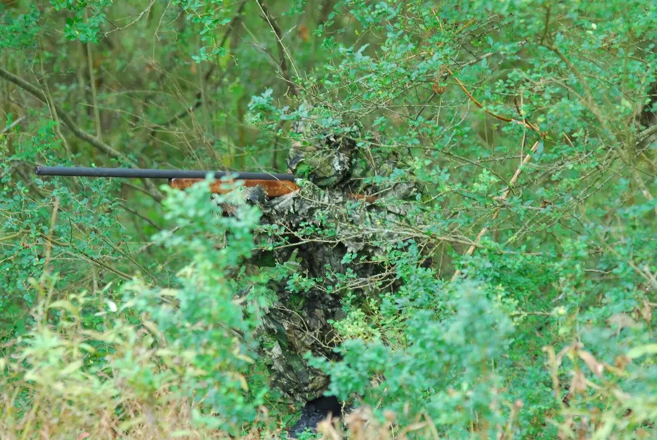 A camouflage-patterned hunting rifle with a black barrel and wooden stock, partially obscured by dense green foliage.