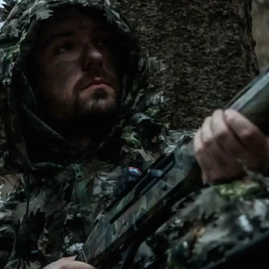 A man in a camouflage hooded jacket and matching camo pants, holding an assault rifle with black stock and barrel.