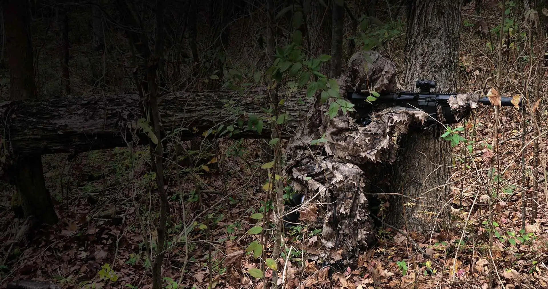 A person in a full-body ghillie suit with leafy camouflage, blending into the forest surroundings.